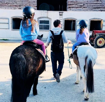 Deux enfants avec casque à cheval sur des poneys guidés par un adulte dans la cour du Manège de Meyrin