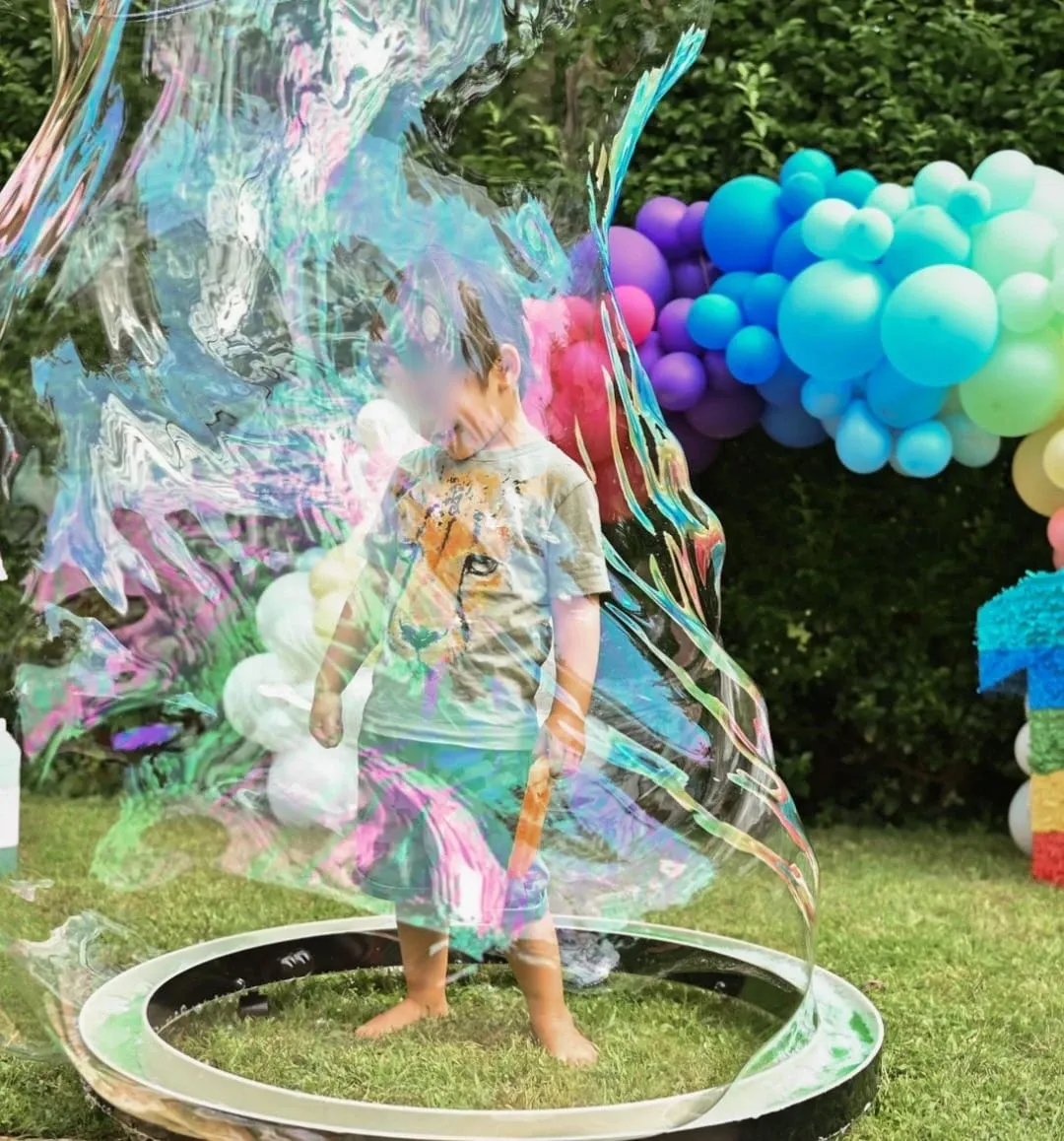 Enfant entouré d'une immense bulle de savon irisée lors du Natalya Bubble Show, décoration de ballons colorés en arrière-plan.