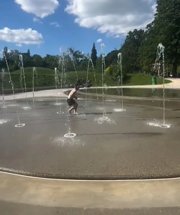 Enfant jouant sous les jets d'eau de la pataugeoire du Bois de la Bâtie, entourée de verdure.