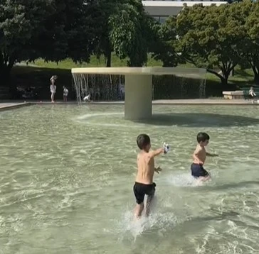 Deux enfants jouant dans la pataugeoire du parc des Franchises à Genève, entourée d'arbres.