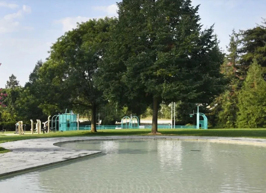 Pataugeoire du parc Trembley avec un grand bassin peu profond entouré de verdure et d'une place de jeux
