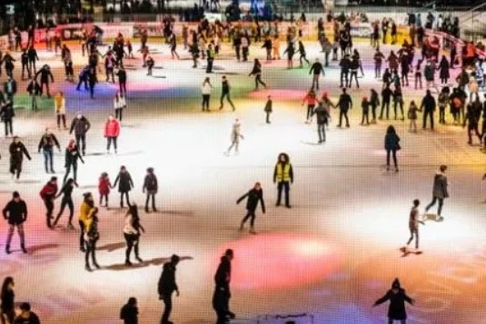 Vue aérienne de la patinoire des Vernets avec de nombreux patineurs sur la glace illuminée de couleurs.