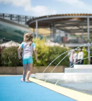 Une petite fille joue près des jets d'eau de la pataugeoire extérieure de la Piscine du Lignon par beau temps.