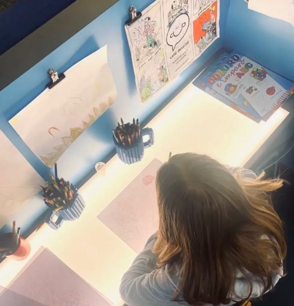 Enfant dessinant à une table lumineuse au Café Dessiné à Genève, entouré de crayons et de BD