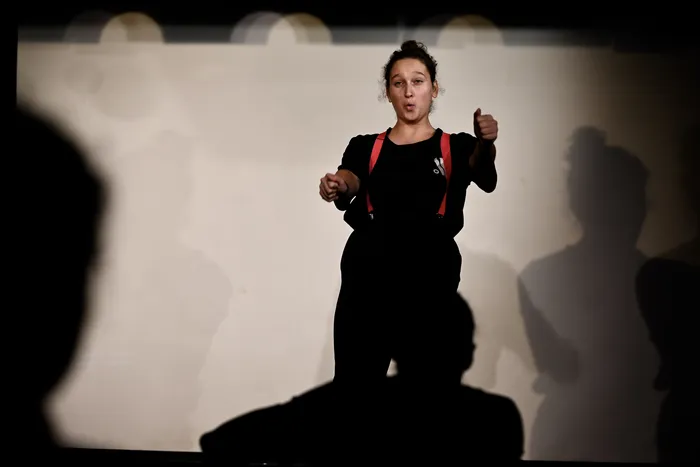 Comédienne en noir à bretelles rouges sur scène, les pouces levés, devant un fond clair avec des silhouettes de spectateurs