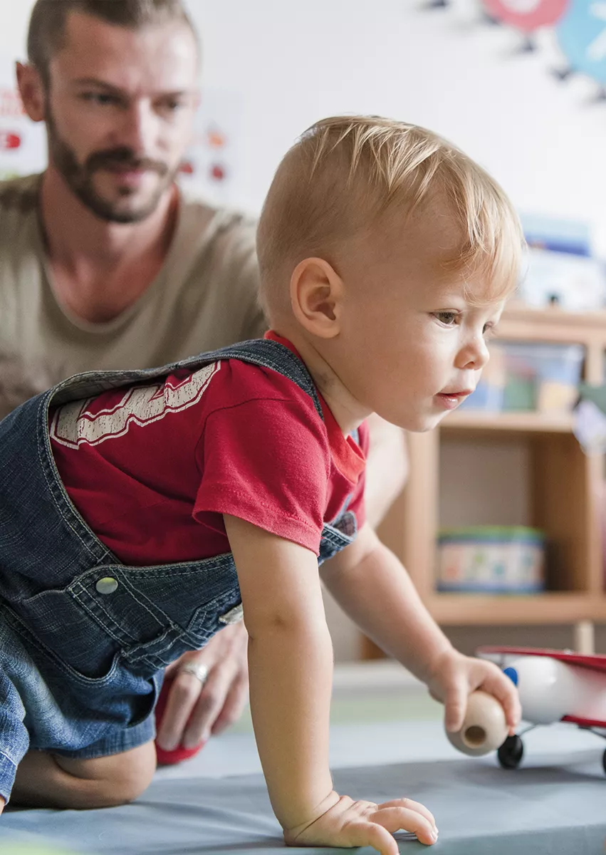 Un jeune enfant à quatre pattes tenant un jouet en bois, accompagné d'un adulte dans un espace d'accueil